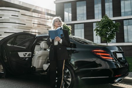 Older professional woman in a suit stands outside a luxury car holding a tablet, with a modern office building in the background.