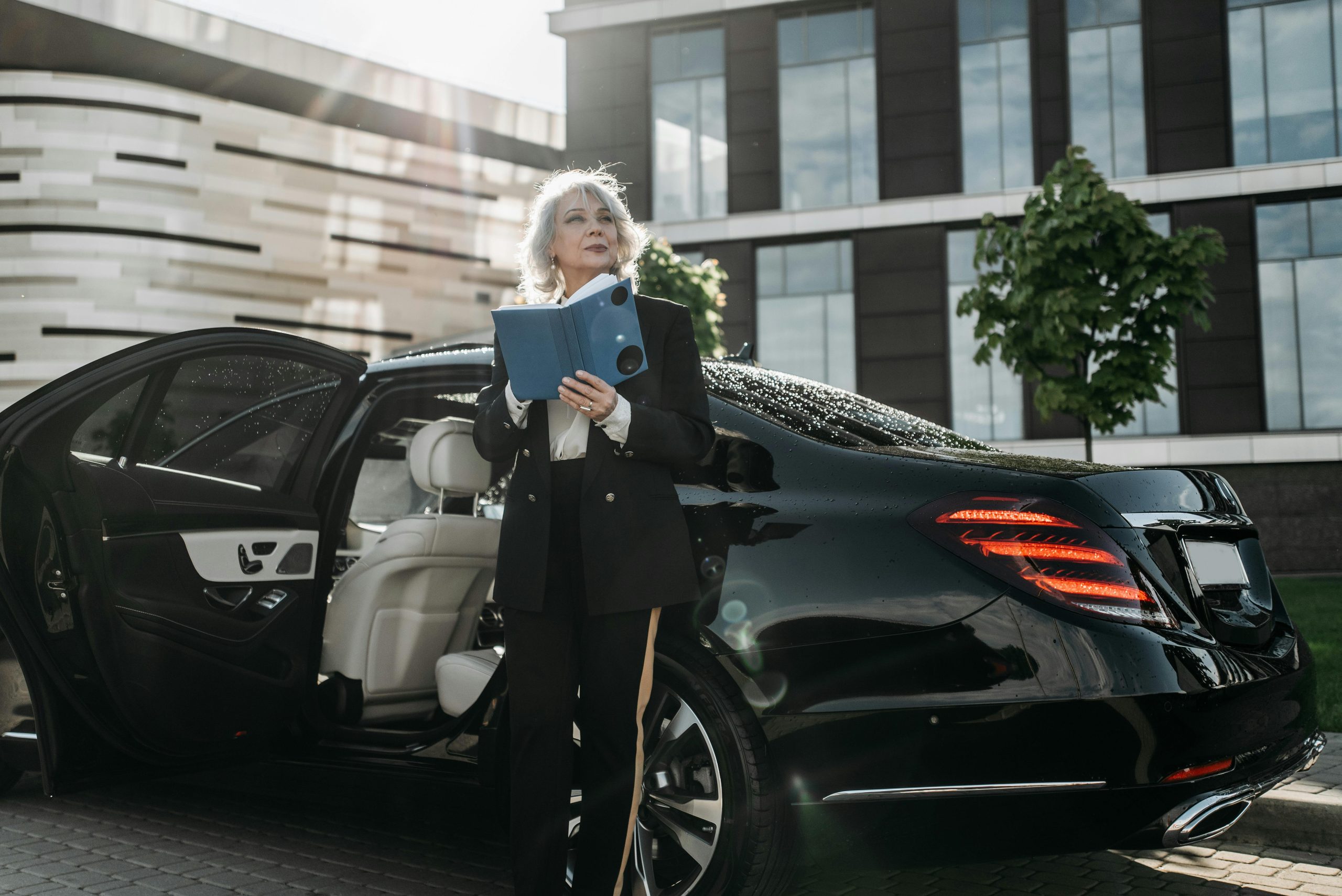 Older professional woman in a suit stands outside a luxury car holding a tablet, with a modern office building in the background.