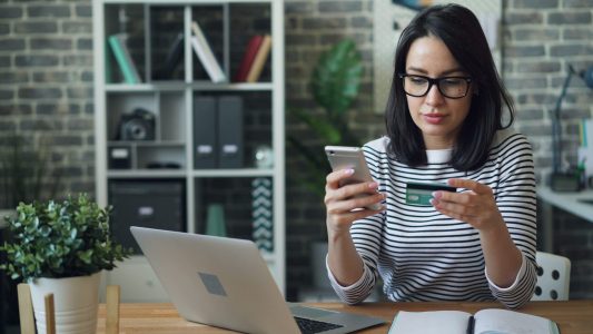 A woman sitting at a table looking at her cell phone and holding credit card; digital payments