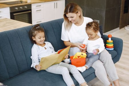 Au Pair sitting on couch and reading young girl a book and holding her baby sister; Childcare Investments