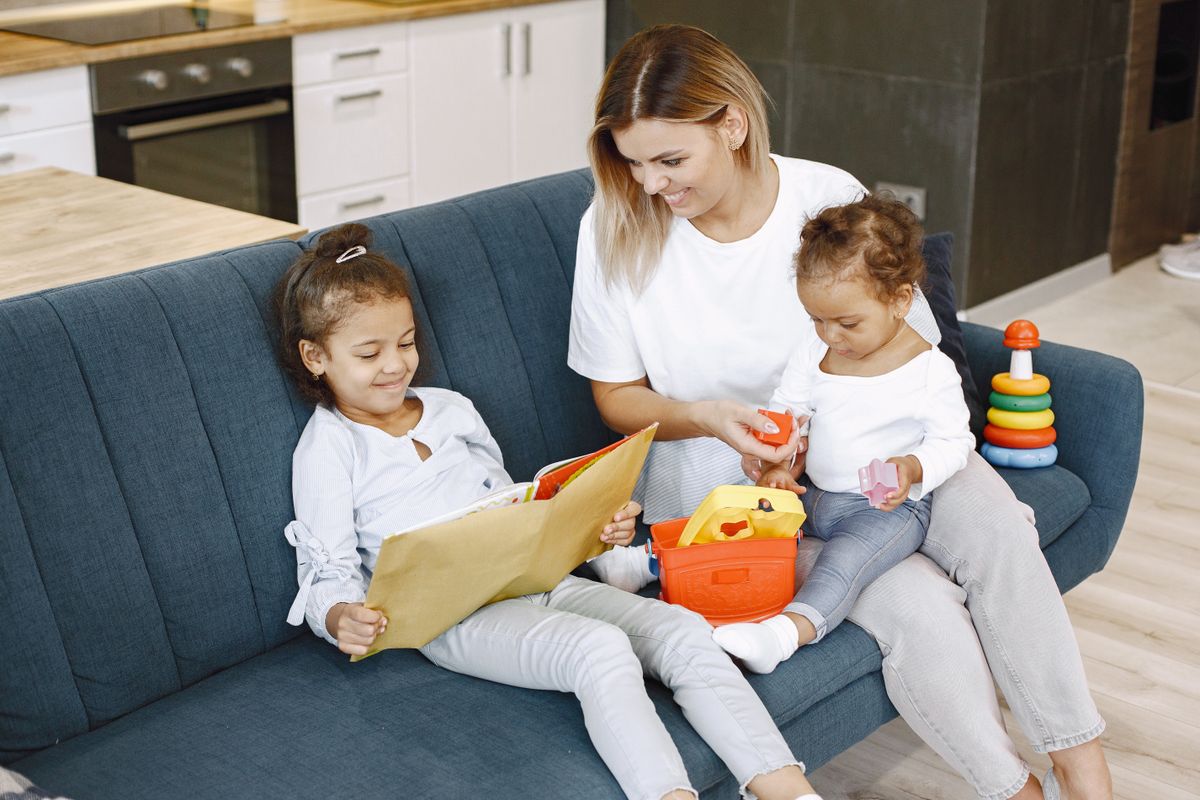 Au Pair sitting on couch and reading young girl a book and holding her baby sister; Childcare Investments