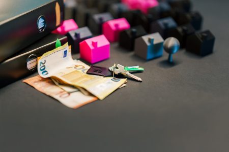 a gray desk with pink and black plastic houses, keys, money; Start Investing in Real Estate Without Buying Property